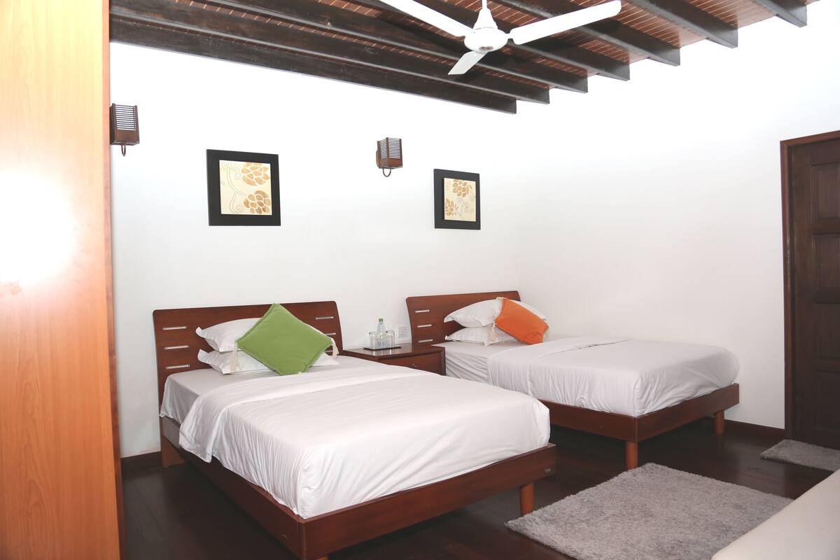 Una habitación con dos camas de madera, ropa de cama blanca, ventilador de techo y cuadros en las paredes.