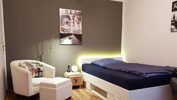 A bedroom with a bed, a chair, a nightstand, and a lamp. - At the gates of Hamburg in the old country (Mittelnkirchen)