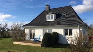 Une maison de plain-pied avec un toit recouvert de tuiles sombres, des murs extérieurs blancs et un petit porche à l’avant, agrémenté de chaises bleues.