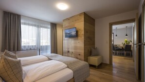 Een moderne slaapkamer met een groot bed, een houten accentmuur, een televisie die erboven is bevestigd, en uitzicht op de stad door dunne gordijnen.