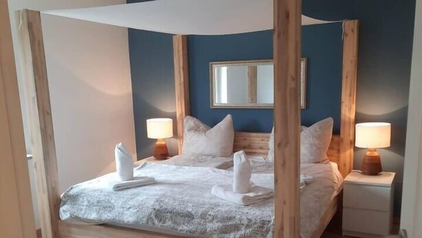 A bedroom with a wooden canopy bed, two bedside tables with lamps, a large mirror on the wall, and a small bench.