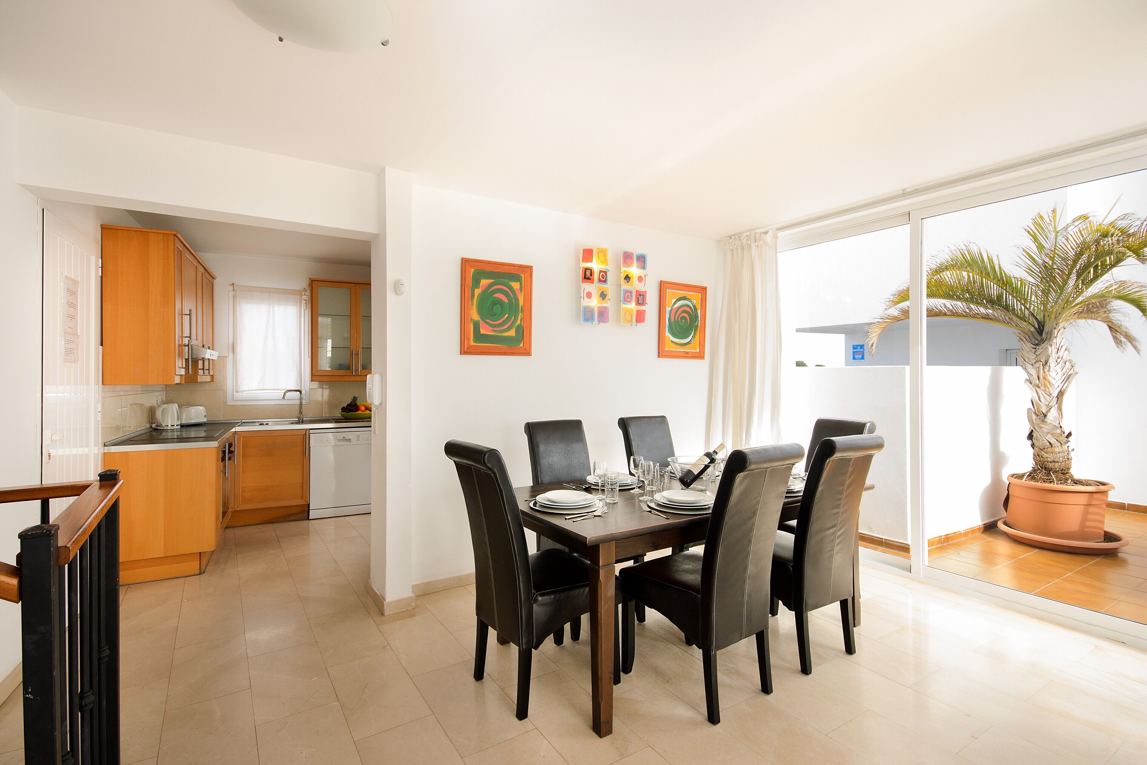 A dining area with a table set for four, a kitchen in the background, and a potted plant by the window.