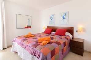 Un letto rifatto con cura, coperto da una coperta colorata, due cuscini rossi e due asciugamani arancioni piegati. Su entrambi i lati del letto, due lampade fissate al muro.