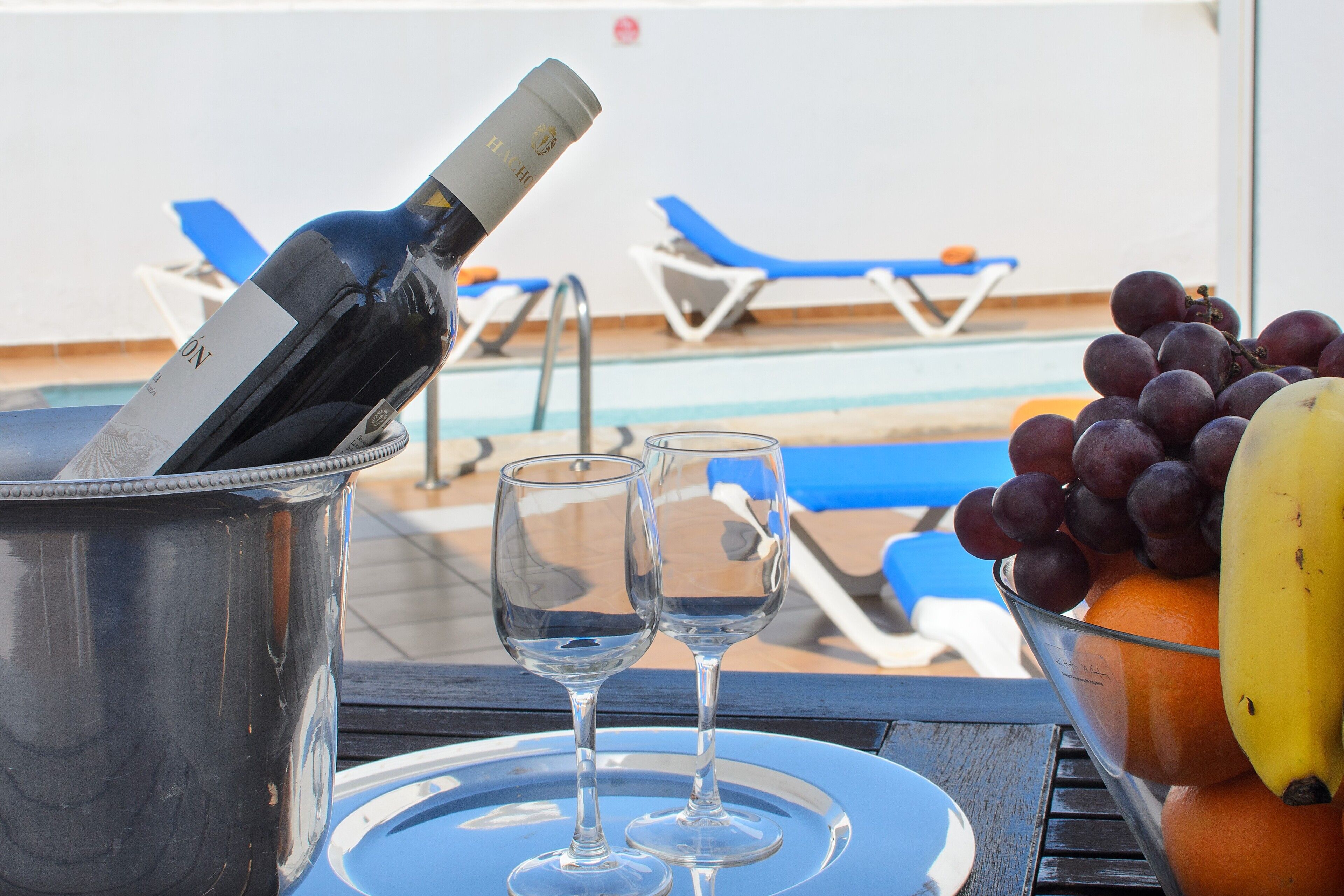 Une table placée au bord de la piscine, avec une bouteille de vin, deux verres et un saladier de fruits.