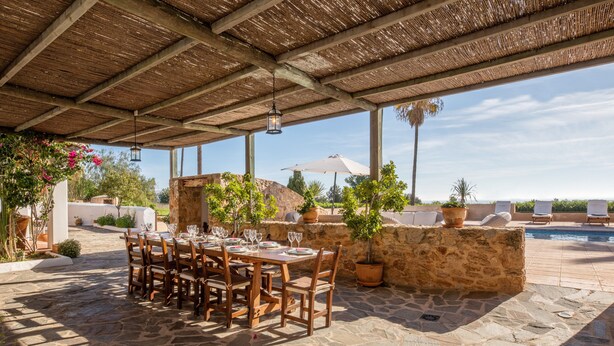 Un patio exterior con suelo de piedra, mesas y sillas de madera, plantas en macetas y una piscina al fondo.