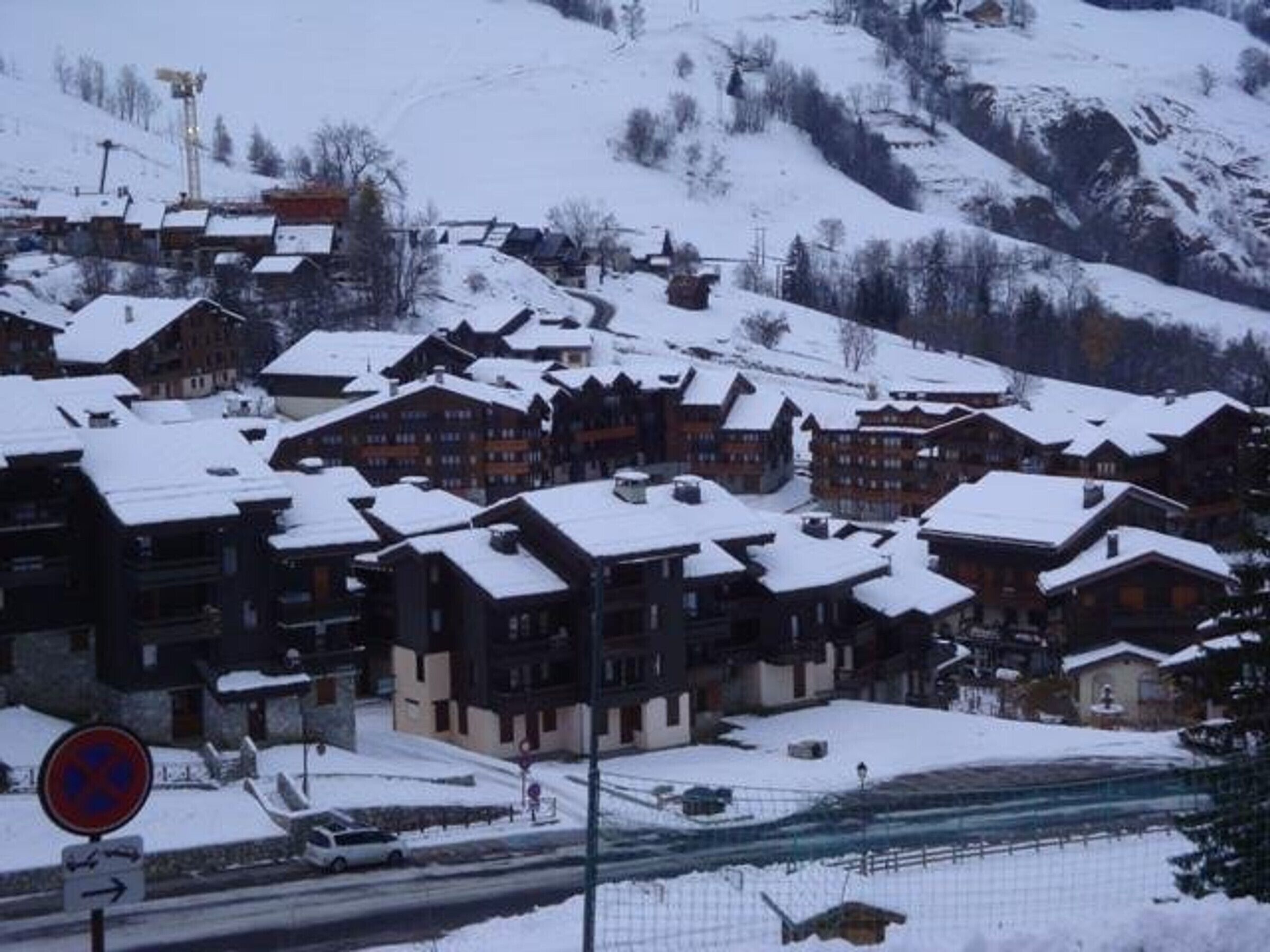 Un pueblo cubierto de nieve con edificios de madera tradicionales, una carretera con señal de prohibido estacionar y montañas al fondo.