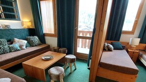 Een gezellige kamer met een houten bank, een kleine eettafel en twee krukken. Er is een raam met gordijnen, een lamp en uitzicht op een balkon.