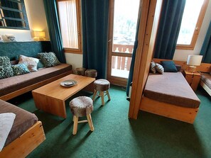 A cozy room with a wooden sofa, a small dining table, and two stools. There is a window with curtains, a lamp, and a view of a balcony. - Cozy studio 4 ppl, ski and shops nearby, pets allowed (Valmorel)