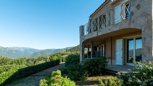 A two-story stone building with a balcony, surrounded by greenery and mountains in the background.