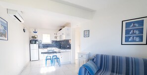A compact living space with a kitchenette, a dining area with blue chairs, and a striped sofa. - APPARTEMENT CARDU  (SAINTE LUCIE DE TALLANO)