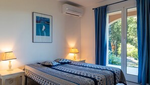 A bedroom with a bed, two bedside lamps, a framed picture on the wall, an air conditioner, and a window with curtains.