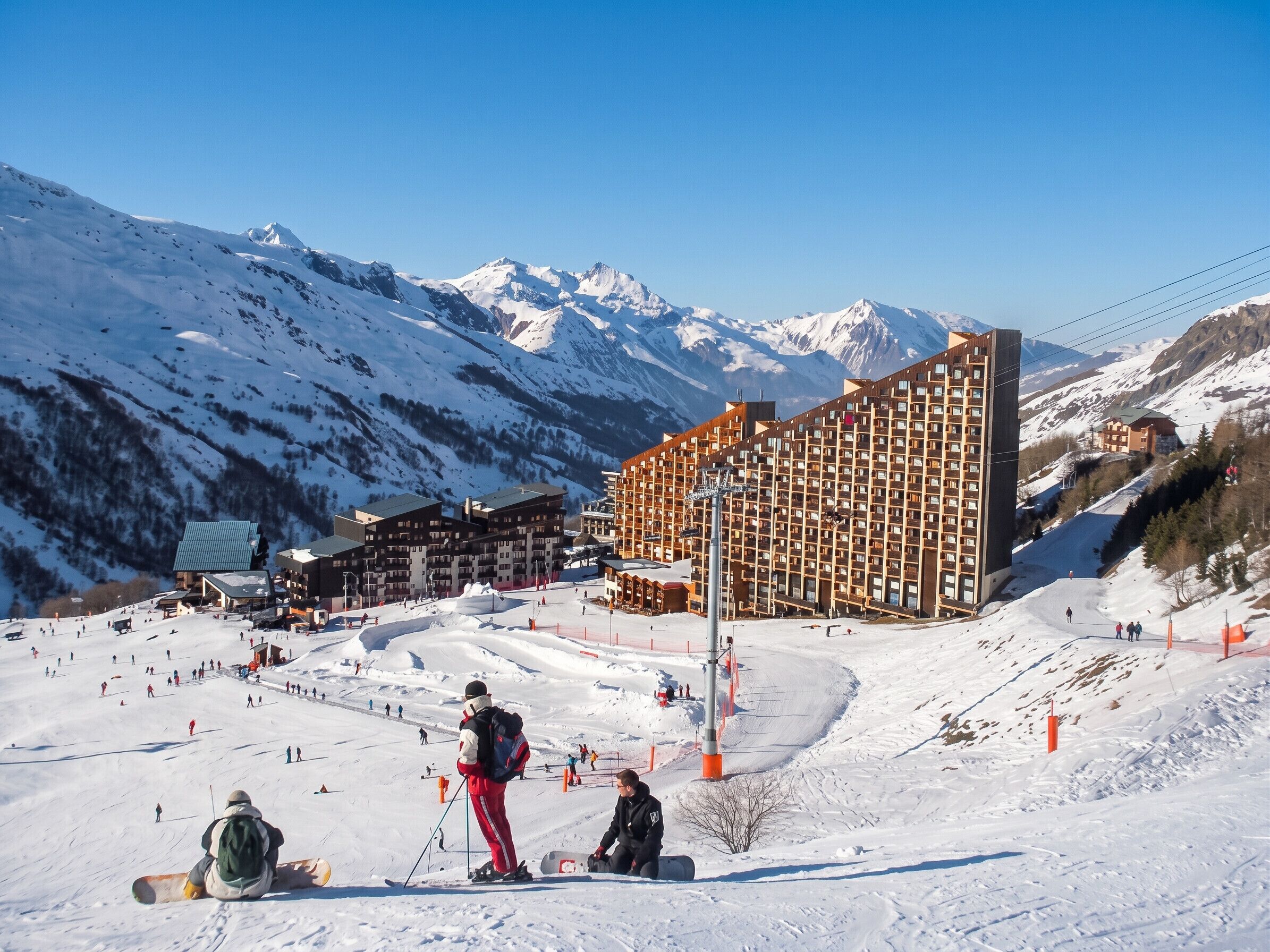 Une station de ski comprenant plusieurs bâtiments, dont un grand hôtel à la façade structurée en forme de grille, des pistes de ski et, en arrière-plan, des montagnes enneigées.