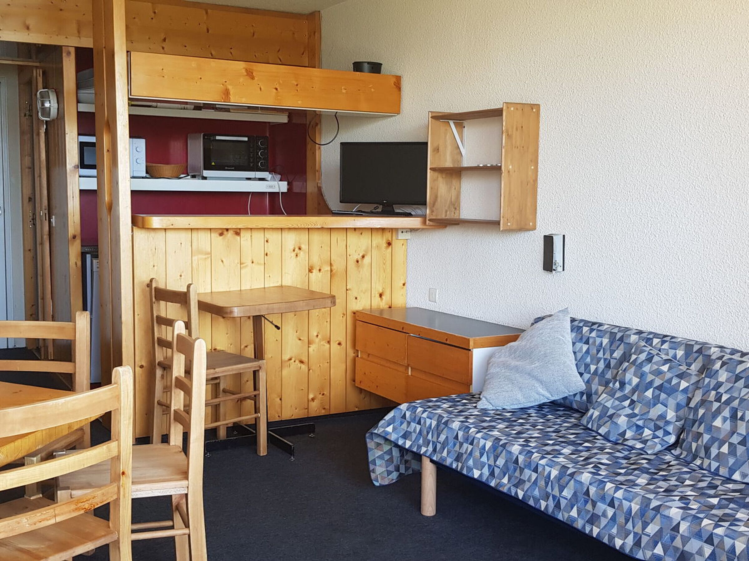 A compact room with a wooden kitchenette, a small dining table, a sofa, and a TV.