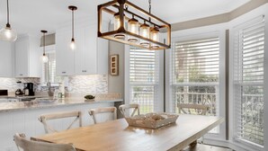 A modern kitchen with white cabinets, a wooden dining table, and a large window with blinds. - Beach Svc⛱️PrivatePool-Gulf Views-Crows Nest-Windancer Amenities-3BR Sun☀️Seeker (Miramar Beach)
