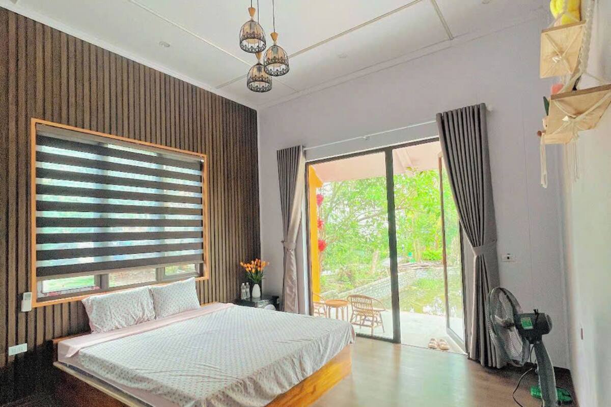 Une chambre moderne avec un grand lit, des lambris en bois sur les murs, un ventilateur de plafond et une vue sur l’extérieur grâce à une porte-fenêtre coulissante.