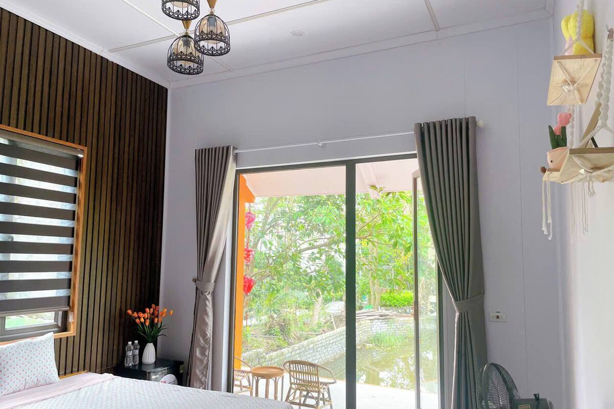 Une chambre à coucher comprenant un lit, une table de chevet, une fenêtre avec des stores, un ventilateur et une vue sur un jardin.