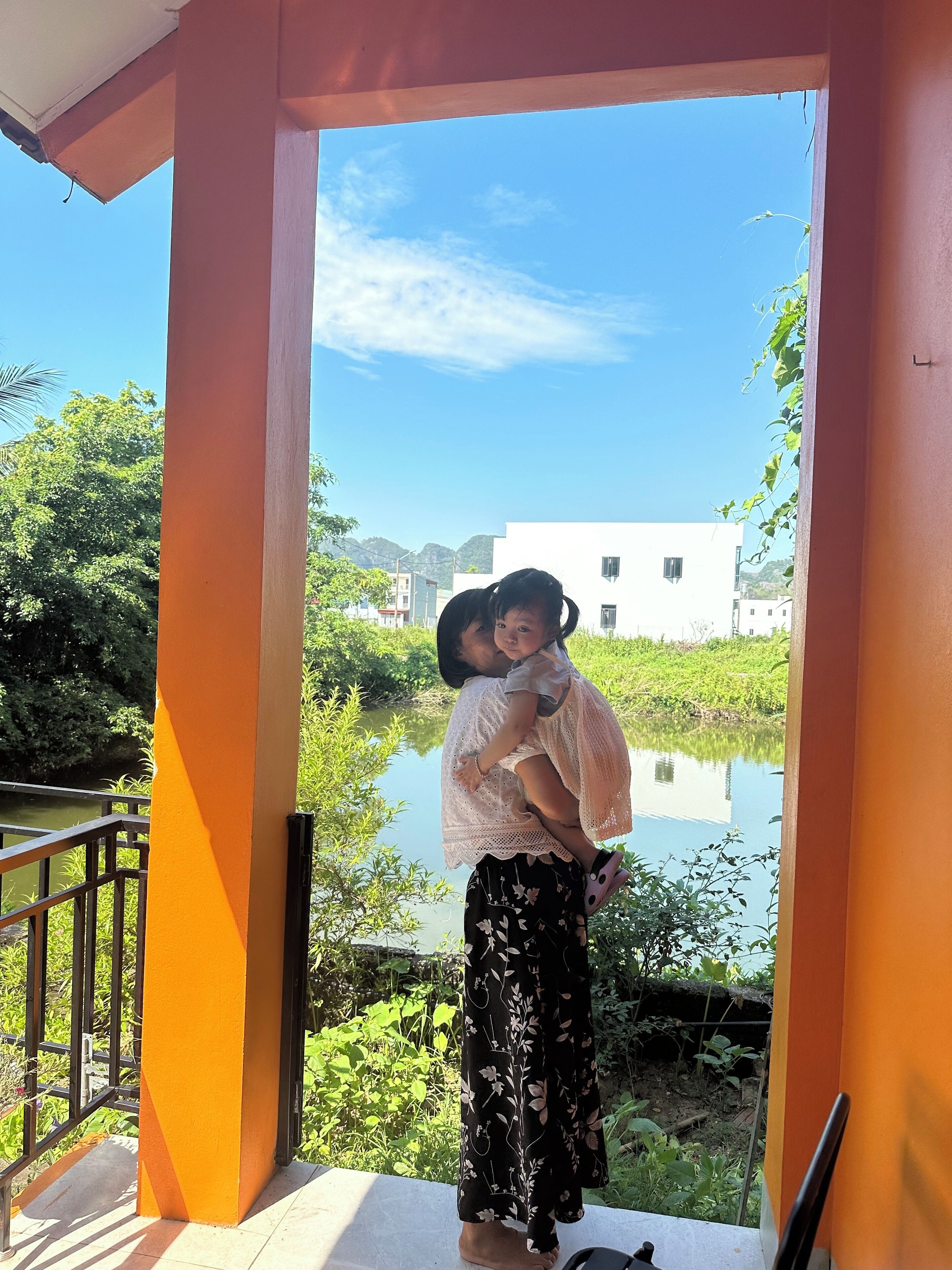 A person carrying a child on their back, standing on a balcony with a view of a water body and greenery.
