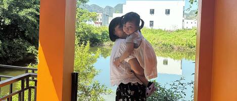 A person carrying a child on their back, standing on a balcony with a view of a water body and greenery.
