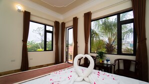Une chambre d’hôtel avec un lit, une serviette pliée en forme de cœur, une fenêtre avec des rideaux, une petite table avec une plante et une chaise.