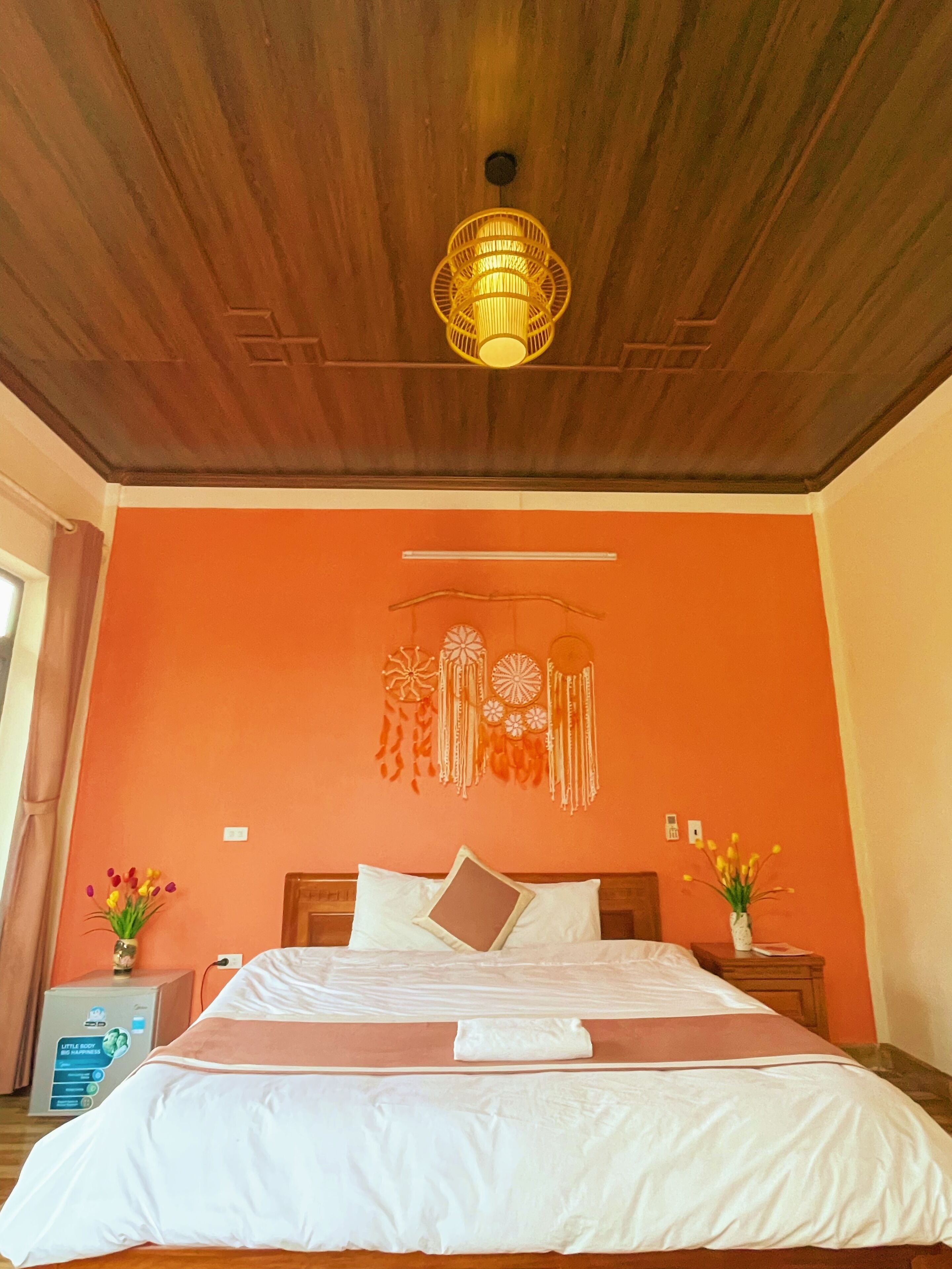 A room with a wooden ceiling, an orange wall, a bed with white bedding, a bedside table with a vase of flowers, and a wall hanging.