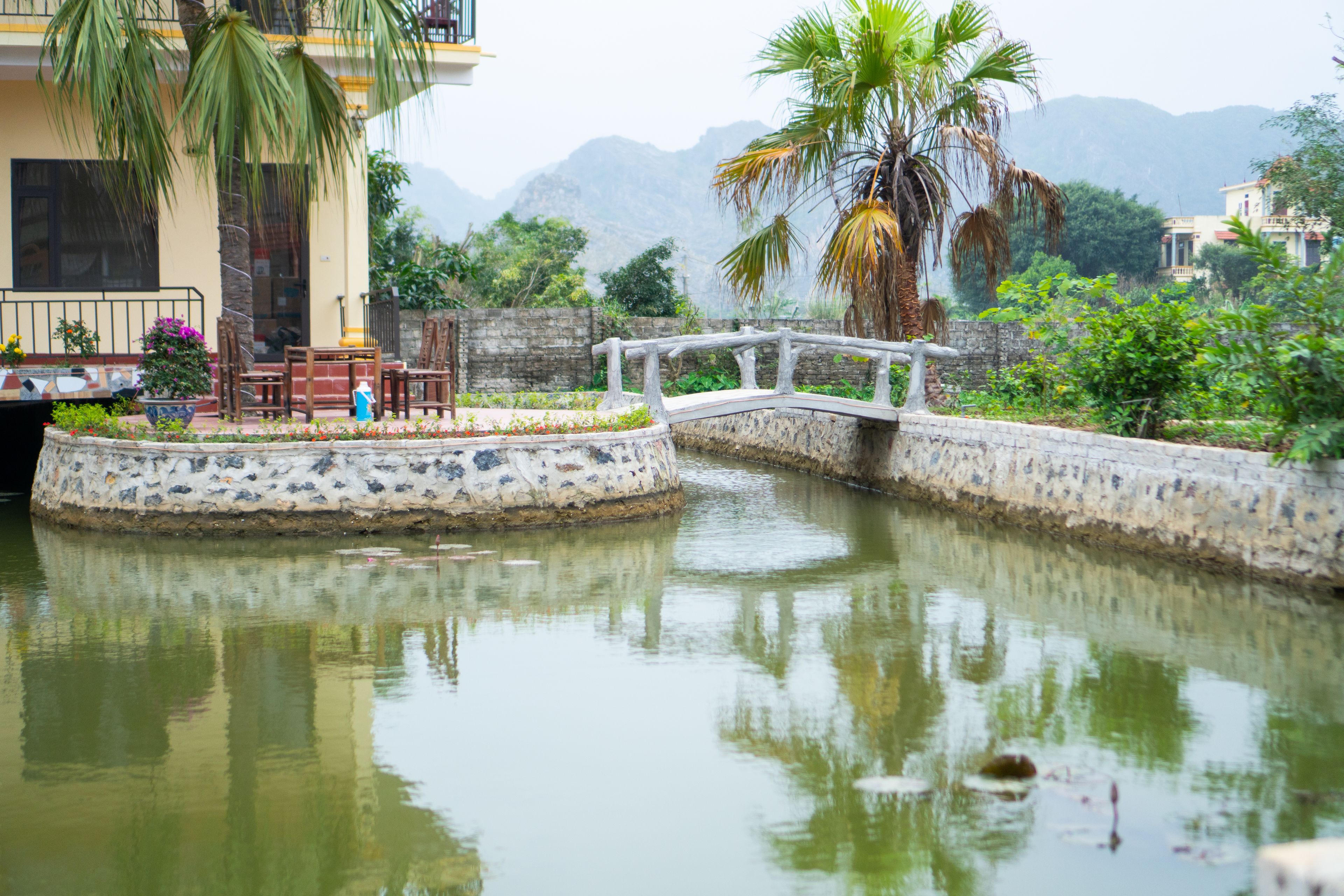 A serene water feature with a bridge, surrounded by lush greenery and a mountainous backdrop.
