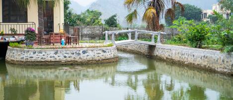 A serene water feature with a bridge, surrounded by lush greenery and a mountainous backdrop.
