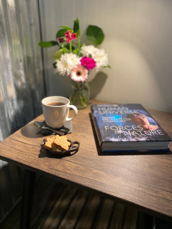 A wooden table with a cup of coffee, a book titled 'Forces of Nature', a small plate with cookies, and a vase with flowers.
