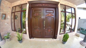 A large, ornate wooden door with intricate carvings, flanked by large windows and potted plants.