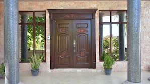 A double wooden door with intricate carvings, flanked by two columns with mosaic patterns, set against a stone wall with large windows.