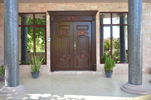 A double wooden door with intricate carvings, flanked by two columns with mosaic patterns, set against a stone wall with large windows.