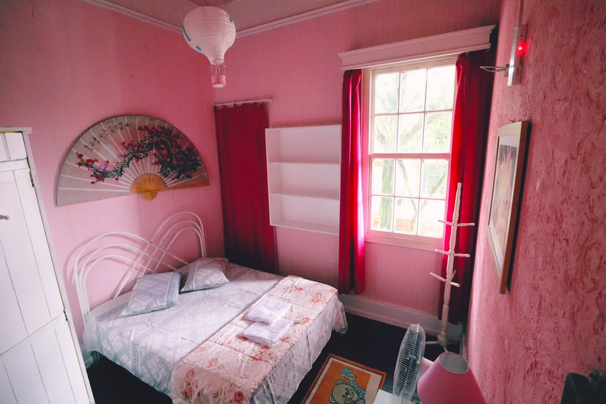 A pink-themed bedroom with a bed, a fan, a window with red curtains, and a wall-mounted heater.