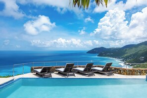 A pool with lounge chairs and a view of the ocean and hills.