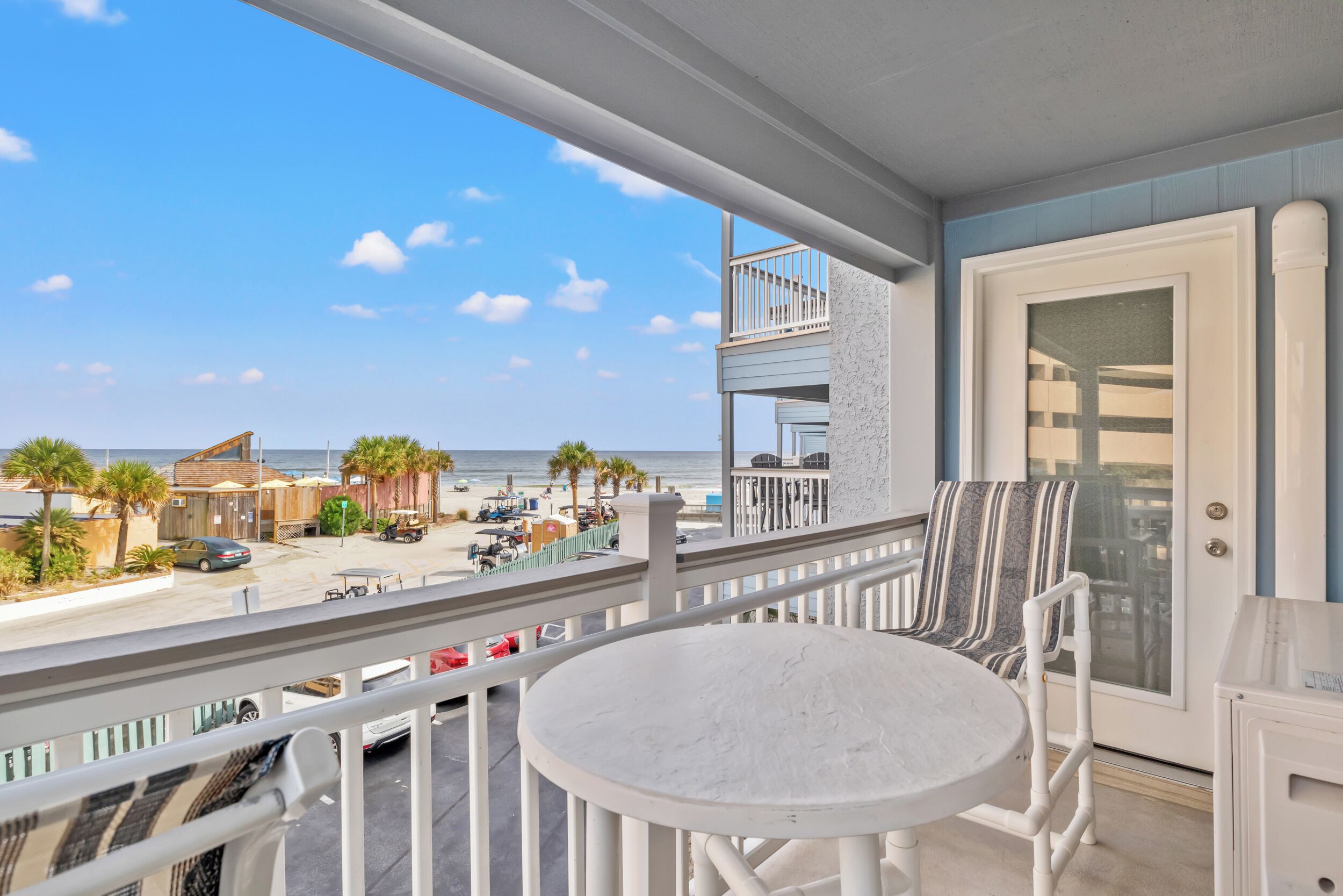 A balcony with a white table and chairs, overlooking a beach with palm trees and a clear sky.