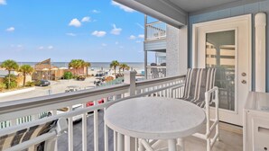 Un balcone con tavolo e sedie bianche, vista su una spiaggia con palme e cielo sereno.