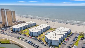 Un hotel frente a la playa con varias construcciones, un estacionamiento y una playa al fondo.