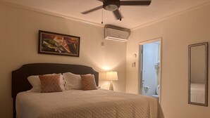 A hotel room with a bed, a ceiling fan, an air conditioning unit, a painting, a lamp, and a bathroom visible through an open door. - Comfy and Secure  One Bedroom Apt- Sleeps 3!! (New Kingston)