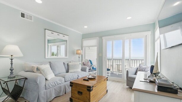Ein modernes Wohnzimmer mit einem hellblauen Sofa, einem Holztisch und einem weißen Schreibtisch mit einem Computer. Ein Balkon bietet Blick auf den Strand.