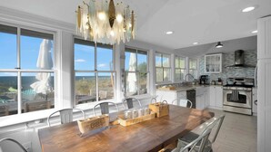 A modern kitchen with large windows, a wooden dining table, and stainless steel appliances.