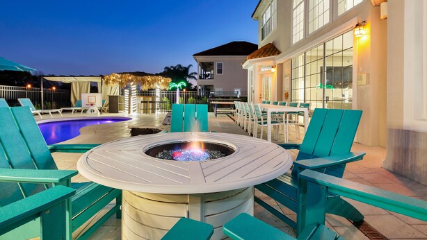 Un espace aménagé au bord de la piscine, avec une table centrale en forme de foyer circulaire entourée de chaises turquoise, une piscine et un bâtiment doté de grandes fenêtres et d’un espace salon extérieur.