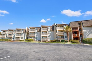 A multi-story residential building with balconies, a parking lot, and a clear blue sky.