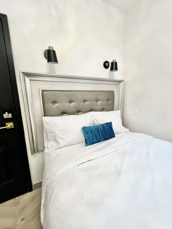 Una cama bien tendida con sábanas blancas y un cojín azul decorativo, un cabecero capitonado y dos mesitas de noche fijadas a la pared con lámparas negras.