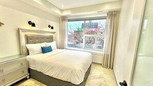 Un dormitorio moderno con una cama grande, una mesita de noche, un aparador y una ventana con vistas a edificios.