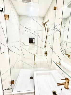Un baño moderno con paredes de mármol blanco, cabina de ducha de vidrio y lavamanos blanco con grifo dorado.