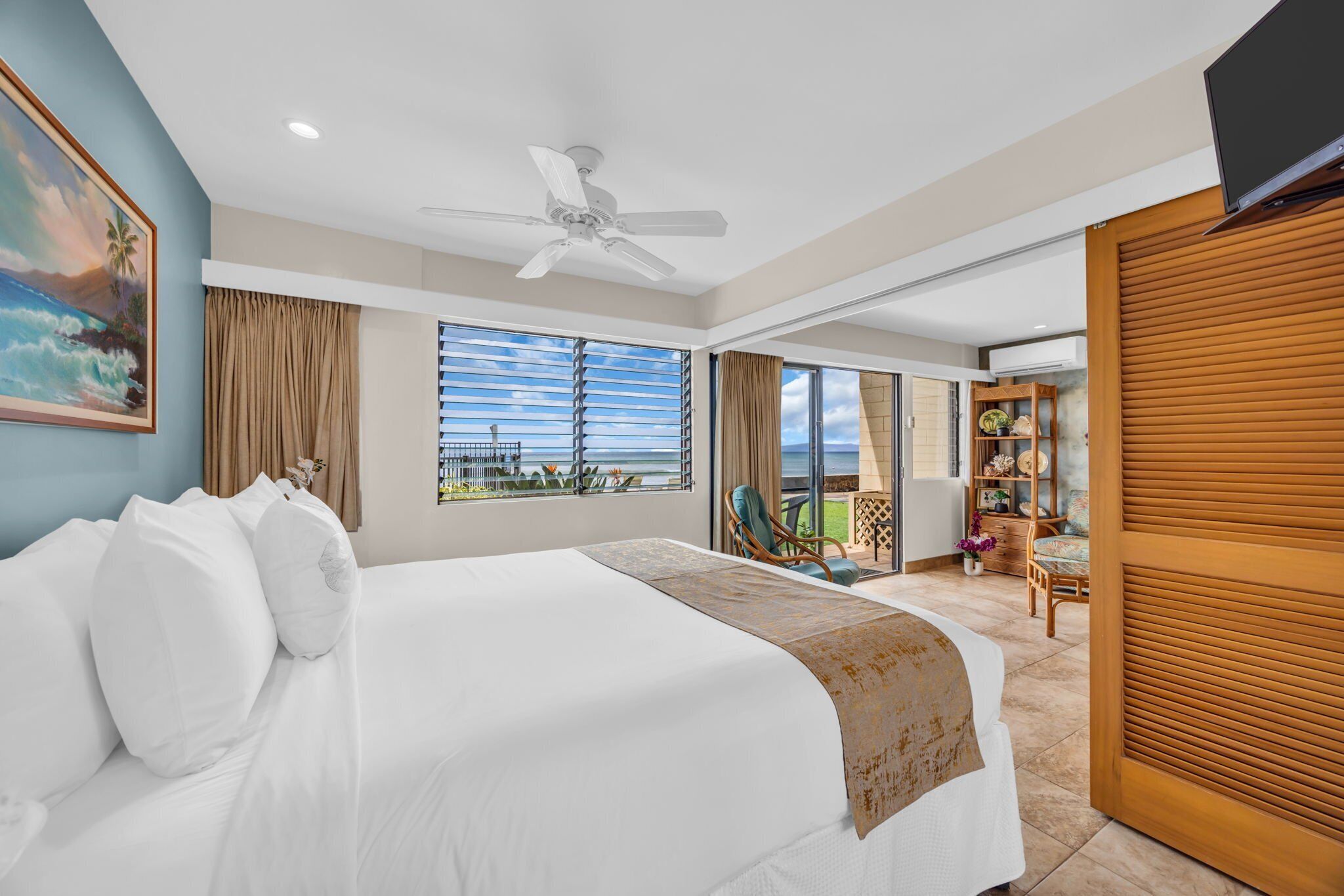 Een hotelkamer met een groot bed, een plafondventilator, een balkon met uitzicht op de oceaan en een aan de muur bevestigde televisie.
