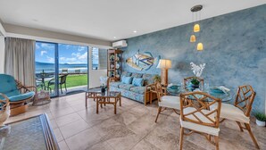Een ruime woonkamer met een grote schuifdeur naar een balkon met uitzicht op zee. Er is een bank, een eettafel met stoelen en een decoratief wandobject.