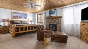 A cozy room with a wooden ceiling, a bed, a leather armchair, a small ottoman, a wooden dresser, a television, and a fireplace.