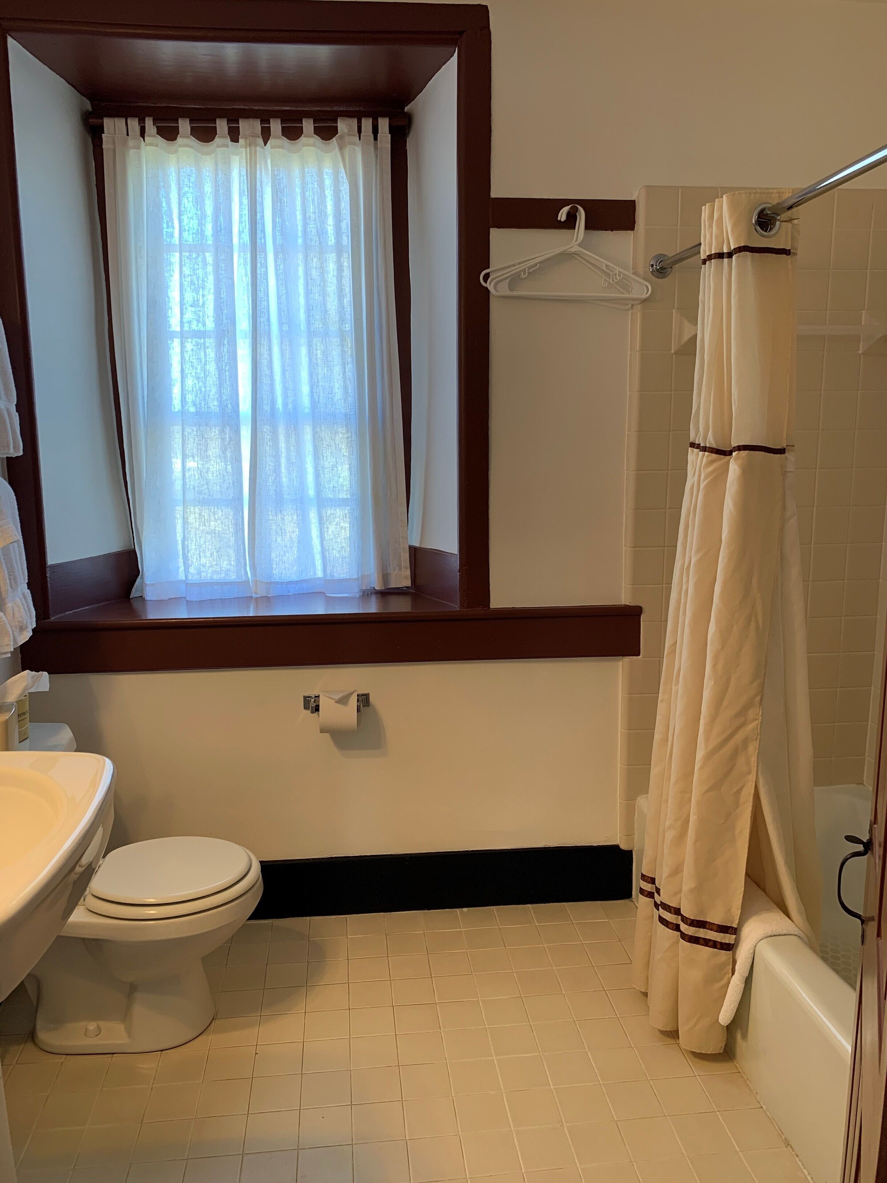 A bathroom with a toilet, sink, window with curtains, and a shower with a curtain.