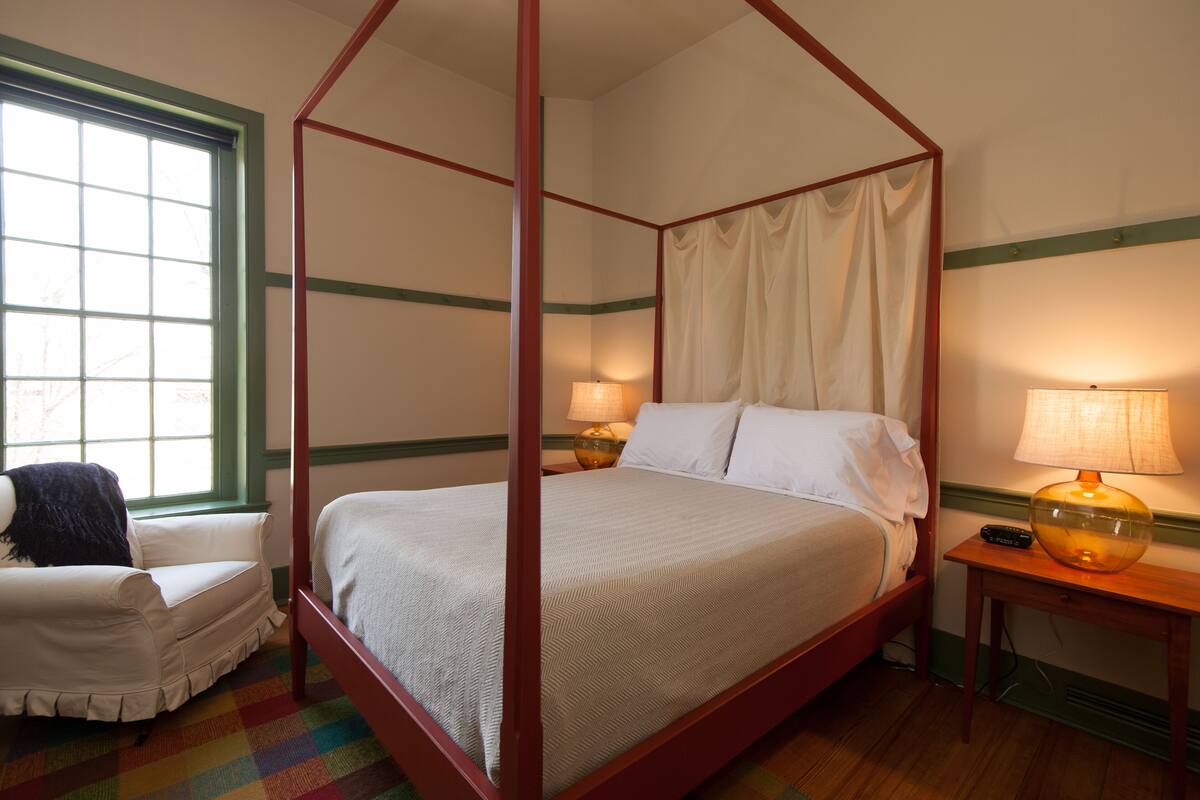 A four-poster bed with a canopy, a bedside table with a lamp, a chair, and a window with curtains.