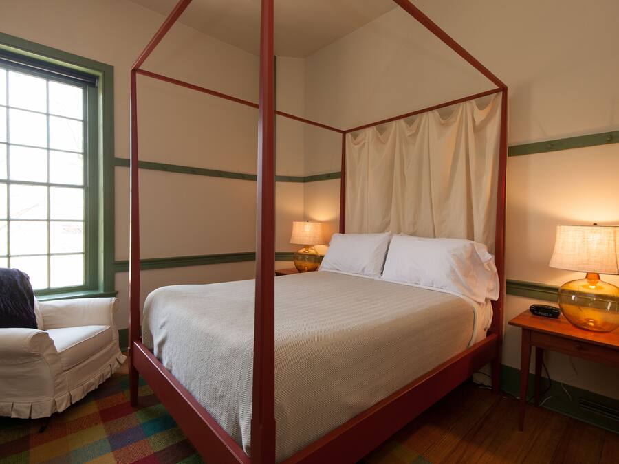 Uma cama de dossel com cortina, uma mesa de cabeceira com abajur, uma cadeira e uma janela com cortinas.