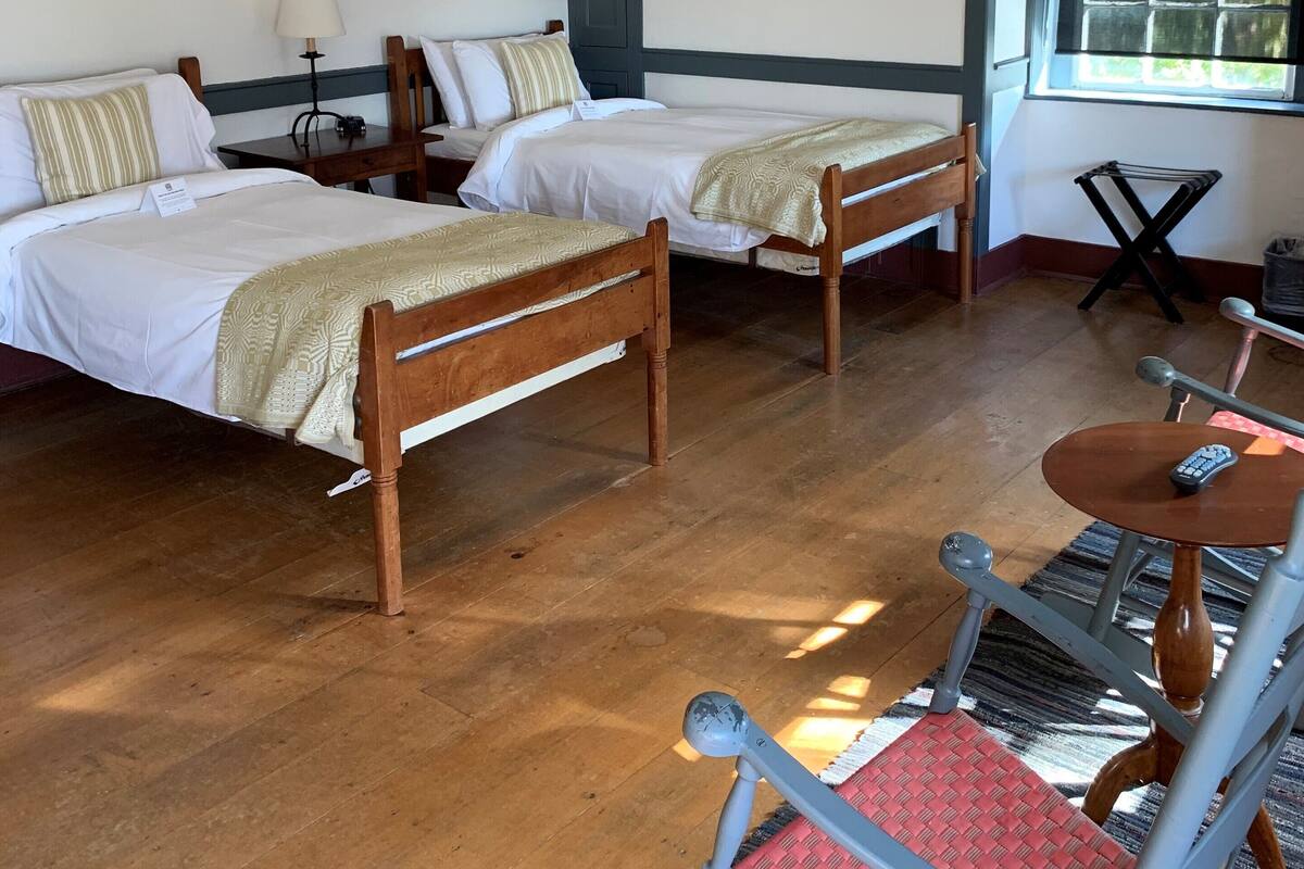 A room with two wooden beds, a small table with a lamp, a chair, and a window with a view of trees.